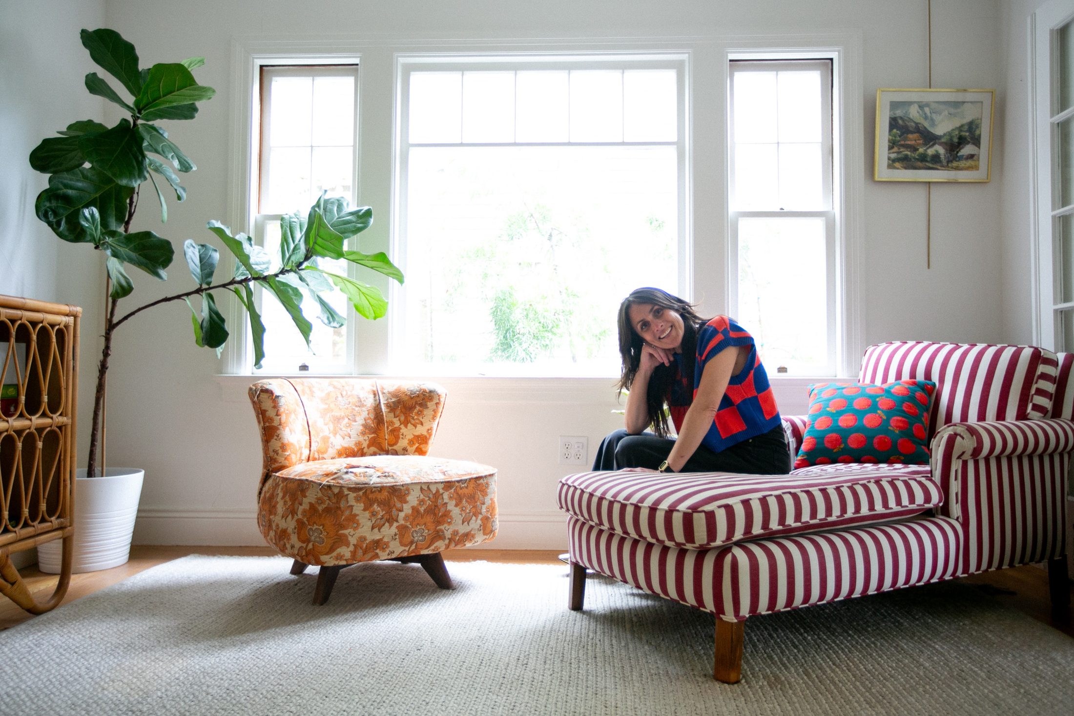 Laura of Tannat Interiors smiling on a red-and-white striped chaise in a bright, plant-filled room